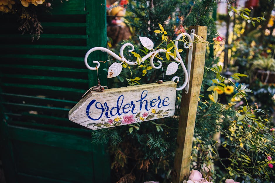 A rustic wooden garden sign with the words 'Order here' painted in blue cursive, hanging from a decorative white metal bracket. The sign is surrounded by vibrant greenery and flowering plants, includi