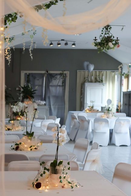 An elegantly decorated interior for a wedding venue, featuring long banquet tables draped with white tablecloths and adorned with floral arrangements. The tables are decorated with delicate white orch