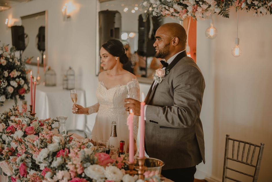 A bride and groom stand at a table decorated with an elaborate floral arrangement featuring pink, white, and blush roses, peonies, and greenery. The bride, with dark hair styled in an elegant updo, we