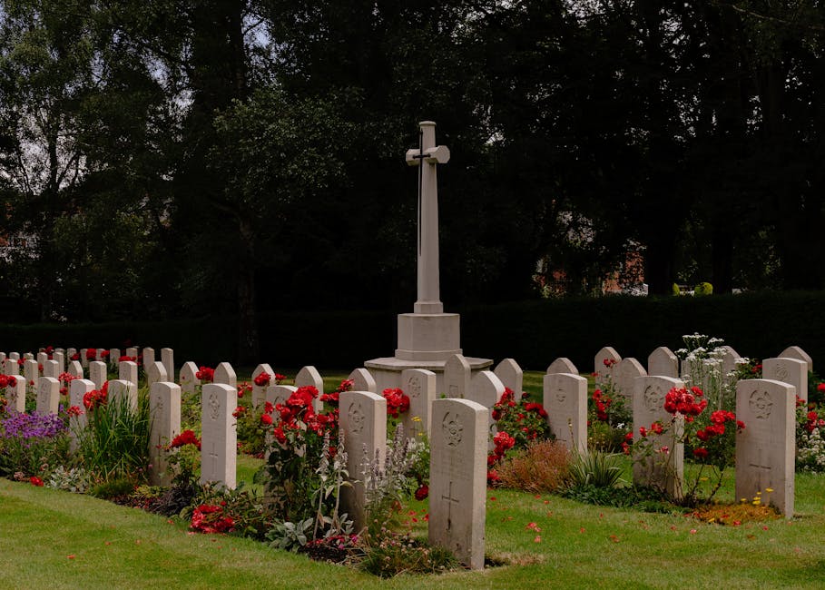 A memorial garden featuring a central white cross monument surrounded by neatly arranged white gravestones, with vibrant red carnations and other colorful flowers planted along the borders. The flower