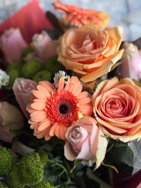 A close-up of a floral arrangement featuring peach-colored roses with soft, layered petals, a single vibrant pink gerbera daisy with a dark center, and clusters of small, green, round buds. The flower
