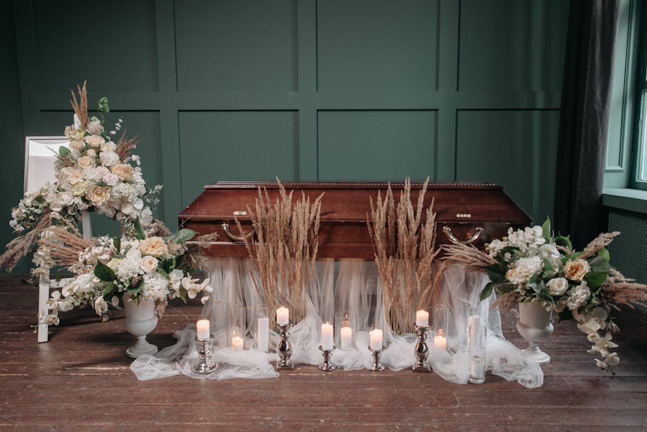 A floral arrangement for a funeral display featuring two large white vases filled with cream and white roses, hydrangeas, and greenery, complemented by tall arrangements of dried pampas grass and whea