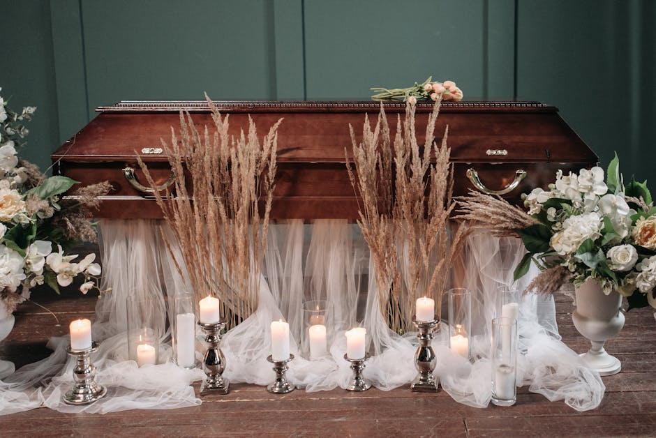 A funeral setting featuring a polished wooden casket as the central focus, surrounded by elegant floral arrangements and candles. The casket is partially draped with sheer white fabric, complemented b