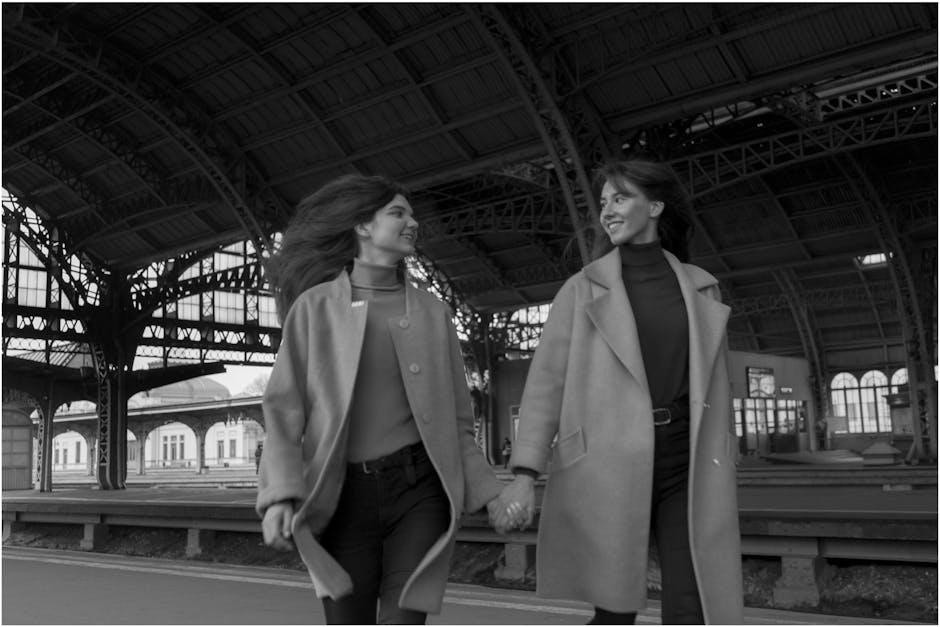 Two women holding hands and walking through a train station with a large, arched metal roof structure and multiple windows in the background. Both women are dressed in stylish, winter-appropriate coat