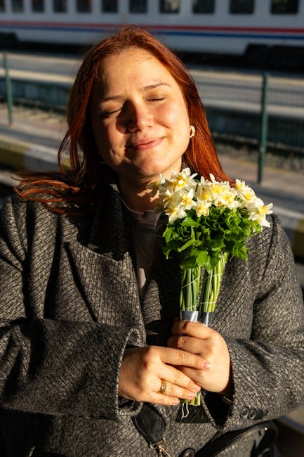 A woman with red hair, her eyes closed and a gentle smile on her face, holding a bouquet of fresh flowers outdoors during daylight. The bouquet features yellow and white daffodils with green foliage, 
