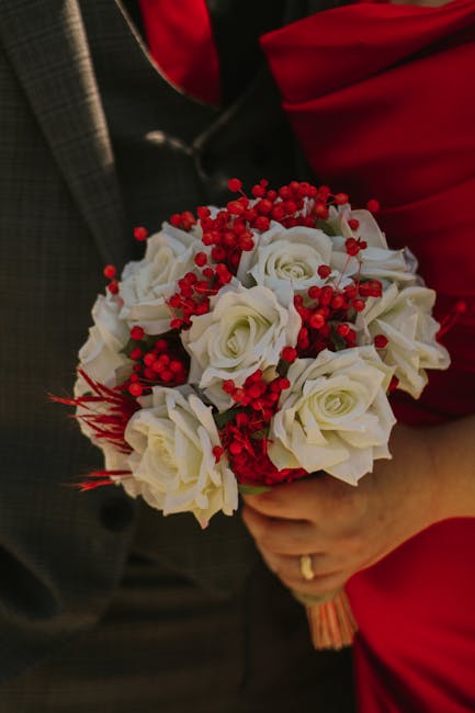 A floral arrangement featuring a bouquet of white roses accented with bright red berries and small red flowers, held by a person dressed in red clothing. The roses have smooth petals and fresh green s