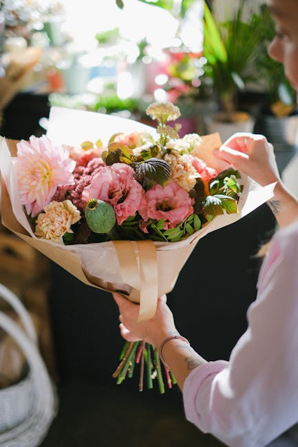 A person holding a large, freshly arranged bouquet of floral gifts featuring pink peonies, cream and white carnations, and delicate purple and yellow filler flowers, wrapped in light brown kraft paper