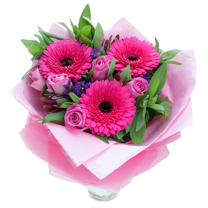 I see this bouquet as a balance between confident colour and soft intimacy: three vibrant fuchsia Gerbera daisies radiate from the centre, their saturated petals and dark centres lending rhythm and focus to the design, while clusters of soft lavender-pink roses nestle close, their tight, velvety buds promising gentle opening over the next days. Intermittent sprigs of purple statice create a delicate, textured contrast - like tiny islands of cool blue-violet set against waves of pink - and the lush green foliage, a mix of broad salal-like leaves and cooler eucalyptus tones, forms a structured yet natural base that holds the composition together. Wrapped for gifting, an inner blanket of pale-pink tissue gives softness to the stems and an outer veil of sheer fuchsia-pink cellophane catches the light with a whispery shimmer; the folds are neat and considered, reflecting careful floristry craft. The lower edge shows the mouth of a clear glass vase, signalling the bouquet's readiness to be placed on a windowsill or coffee table. In the soft afternoon light the petals possess a satiny sheen, the roses release a quiet perfume, and the gerberas offer a faint, green sweetness - a sensory pairing that feels both celebratory and consoling. Perfect for brightening a room after a stroll through Hornchurch Country Park or as a thoughtful gift collected from the town centre, this arrangement honours simple pleasures and neighbourhood gatherings, suited to birthdays, anniversaries, or an uplifting 'just because' moment in Hornchurch and surrounding streets.